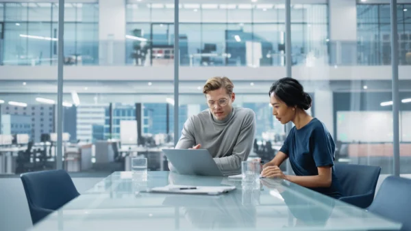 Two colleagues sitting at a desk in a glass-walled modern office.