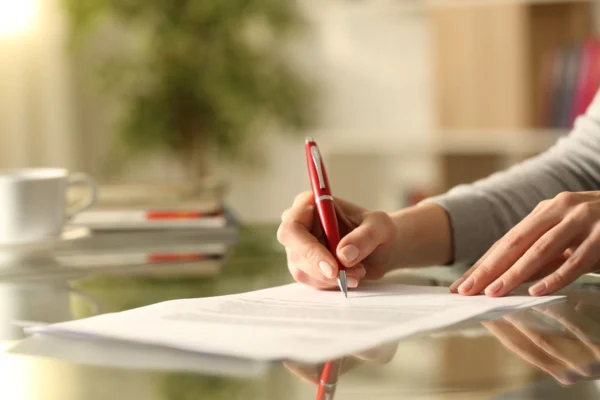 Close-up of someone signing paperwork on a glass table.
