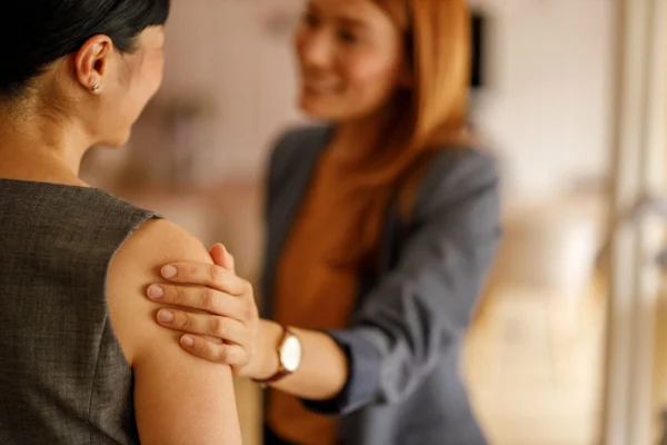 A woman in a blazer offers a supportive gesture by placing a hand on another woman's shoulder.