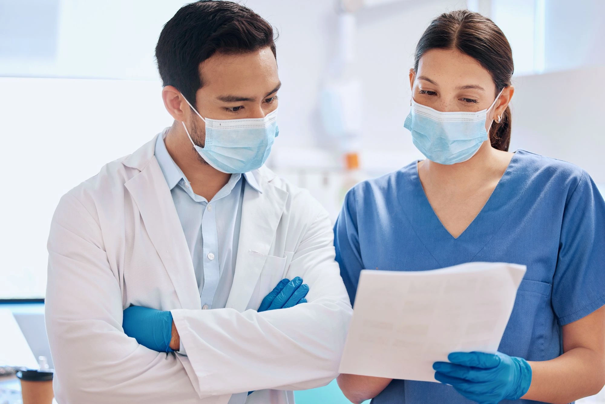 Two medical professionals, wearing masks and blue gloves, review a document together in a clinical setting.