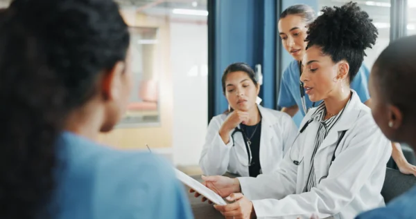 A diverse team of female medical professionals in lab coats and scrubs gathers around a table to discuss a document.