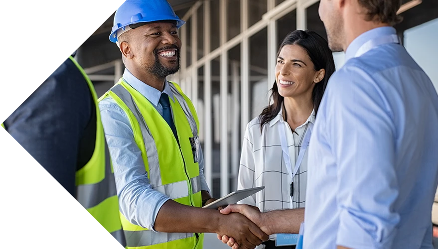 Four professionals, including two construction workers in blue hard hats and yellow safety vests, shake hands and smile during a meeting at a construction site.