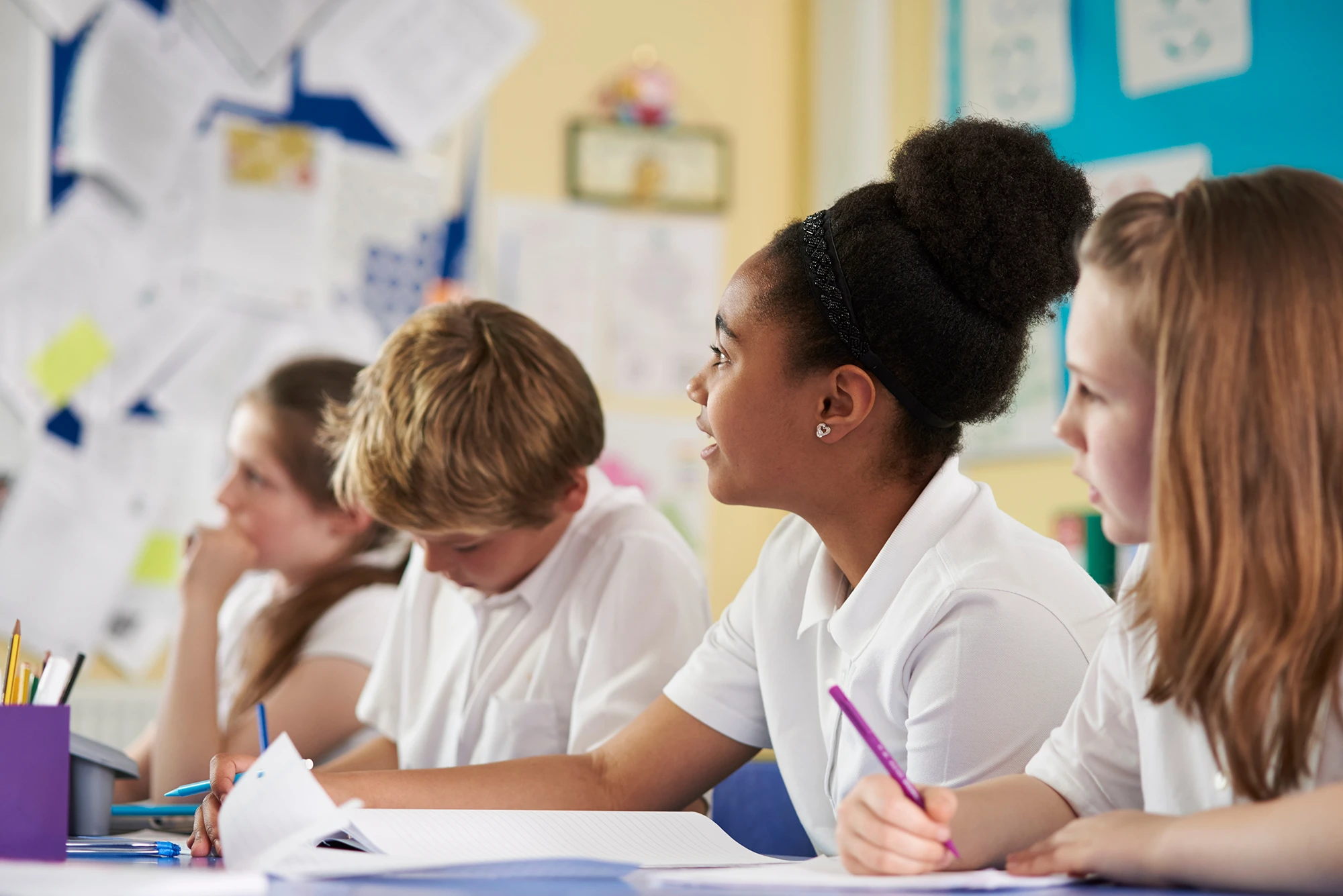 Four engaged primary school students in a classroom writing and listening to teacher.