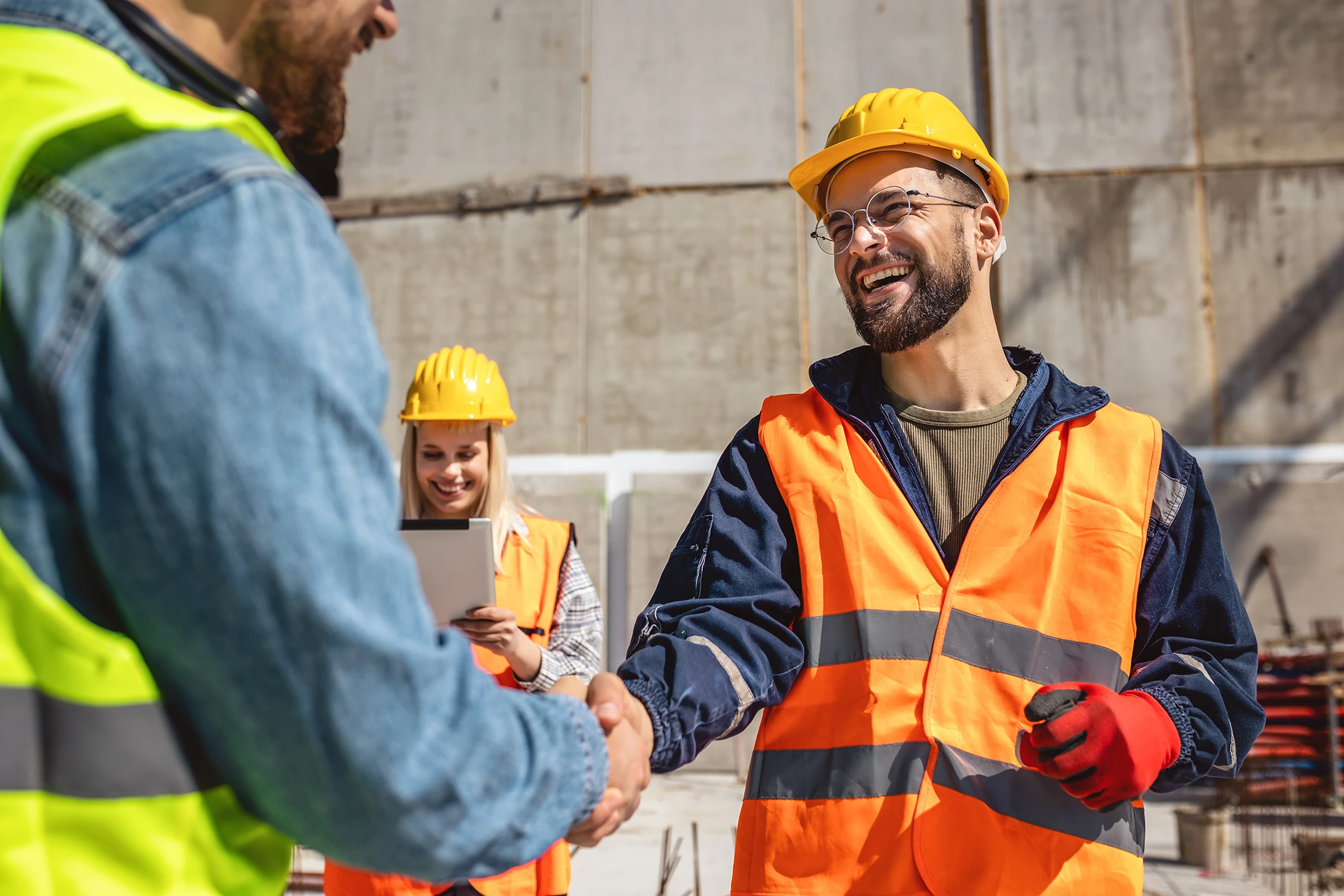 Two construction workers in high-visibility vests and hard hats shaking hands on a job site.