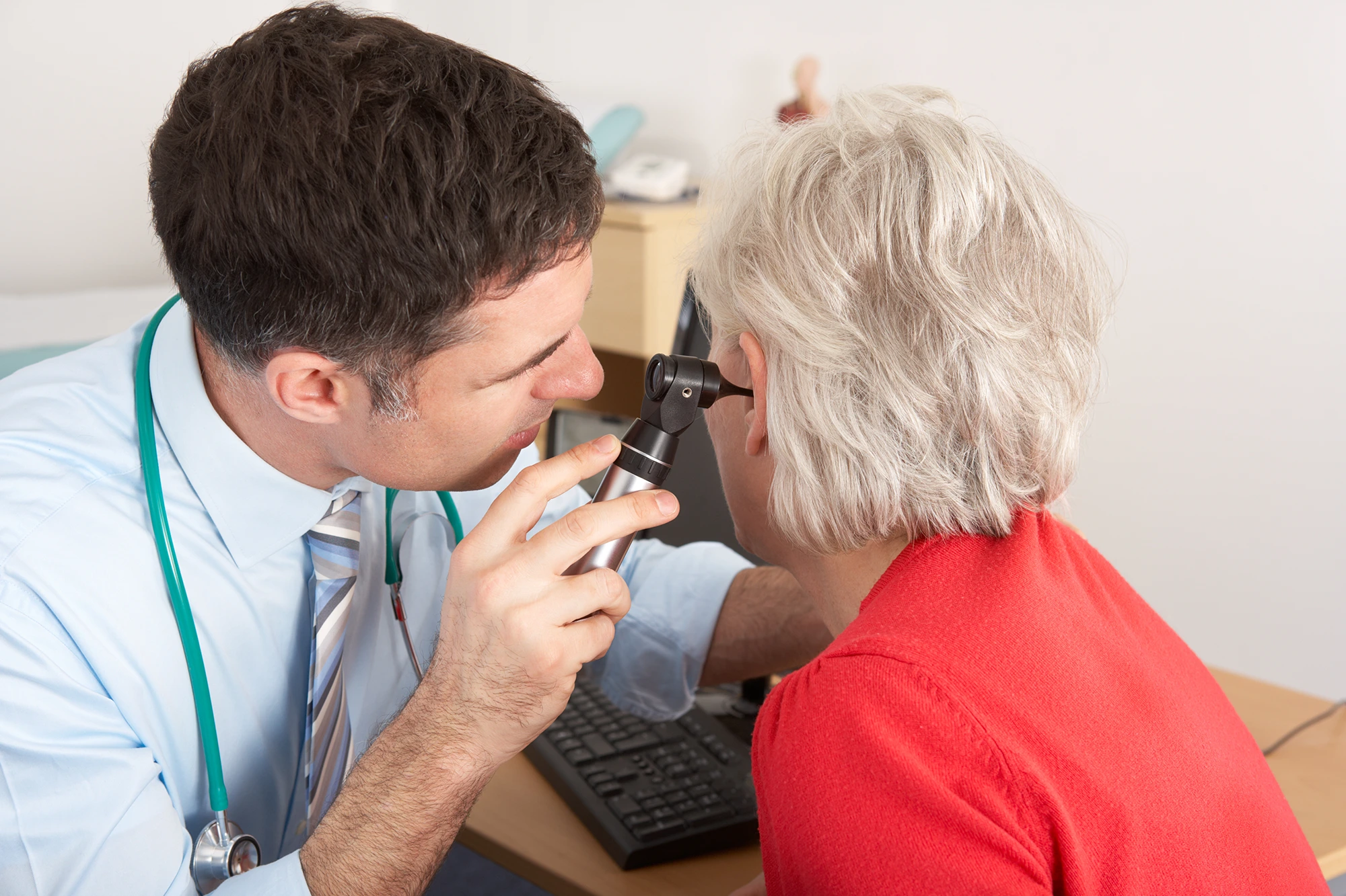 A doctor in a blue shirt is using a black otoscope to look in a female patient’s ear.
