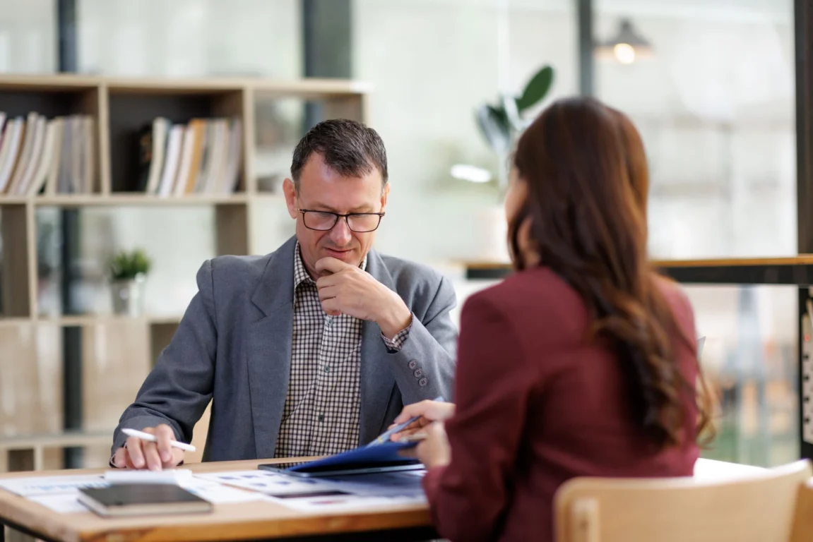 A man in glasses and a suit sits and talks to a colleague at his desk.