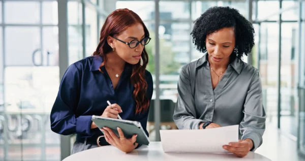 Two employees sat at a round table in an office, one looking at some paperwork, and the other holding a tablet and stylus.