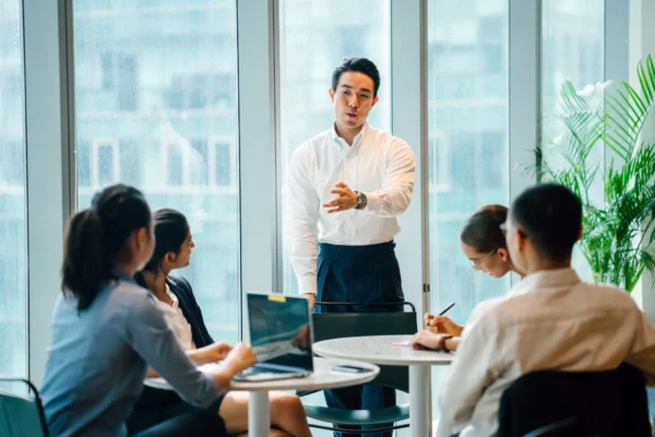 An HR professional providing a training meeting to colleagues in a bright and airy office with floor to ceiling windows.