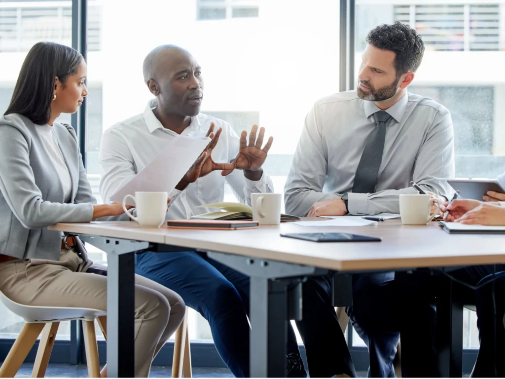 Five office workers sat around square table covered in mugs and paperwork, having a discussion. 
