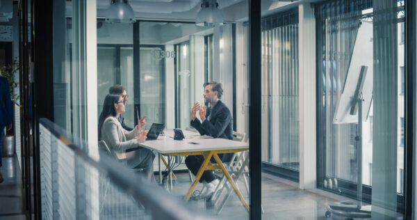 An employee sat across from two businesspeople in a formal discussion across a white table in a glass-walled office. 