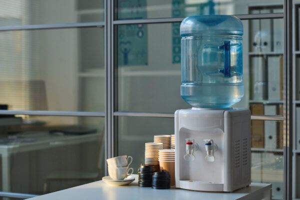 A water cooler on top of a white table, with disposable cups and mugs beside it. 